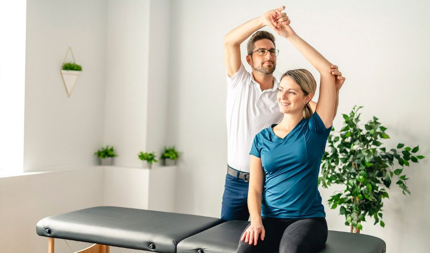 Image of physical therapy with a male physical therapist helping a female patient stretch her arm.