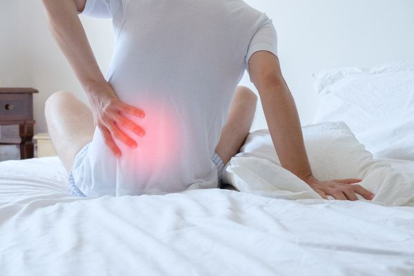 Image of man sitting up in bed and holding his painful back because he needs sciatica treatment.