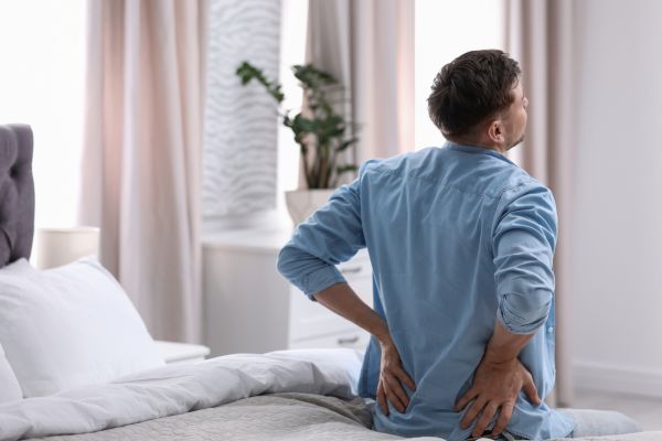 A man sits on the side of a bed, holding his lower back in discomfort