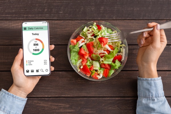 An image of a person eating a salad while counting calories with a smartphone app.