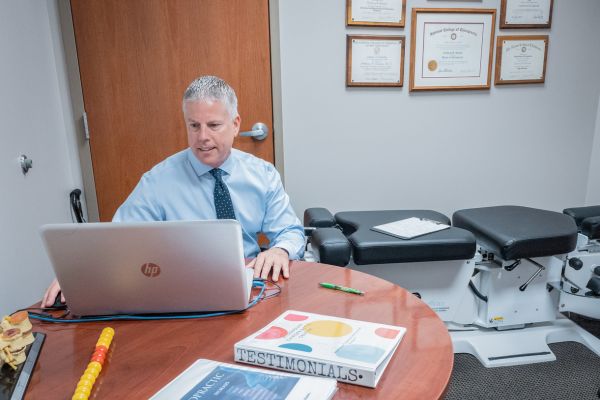 Dr. Galante working at a table with his degrees and certifications framed behind him